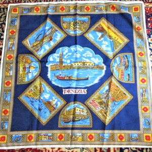 May include: A square, navy blue scarf featuring illustrations of Venice. The center shows a gondola on the water with buildings and a tower. The word "DENEZIA" is printed below. The border has repeating images and a gold and red floral pattern.