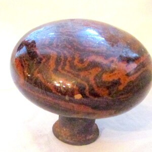 May include: A decorative cabinet knob with a rounded, oval shape. The knob features a swirling pattern of dark brown and reddish-brown hues, with a glossy finish. The base is a darker, muted brown color.
