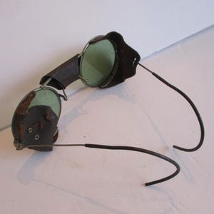 May include: Vintage leather-covered safety goggles with green tinted lenses and a metal frame. The goggles have a flexible metal headband.
