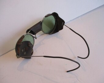 May include: Vintage leather-covered safety goggles with green tinted lenses and a metal frame. The goggles have a flexible metal headband.