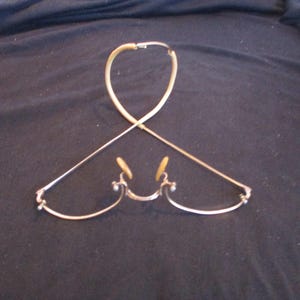 May include: A pair of gold-toned glasses with a flexible, adjustable nose bridge and earpieces. The glasses are folded in half and laid flat on a dark surface.