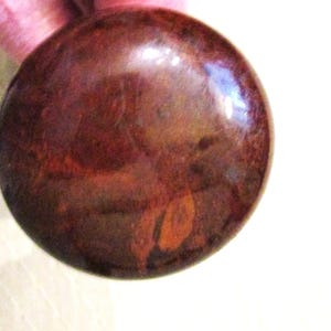 May include: A smooth, circular, polished stone with a deep reddish-brown color and subtle, darker mottled patterns throughout its surface. The object has a glossy, reflective finish that catches light, highlighting its rounded, convex shape.
