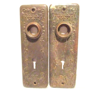 May include: Two antique bronze door plates with ornate floral and scrollwork designs. Each plate features a circular knob, a keyhole, and a small hole at the bottom. The plates have a weathered appearance, suggesting age and use.