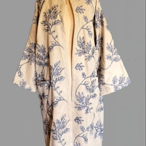 Könnte beinhalten: Beige Kimono-Jacke mit offenem Frontdesign und blauem Blättermuster. Die Jacke hat lange Ärmel und zwei Vordertaschen. Das Kleidungsstück wird auf einer Schaufensterpuppe vor grauem Hintergrund präsentiert.