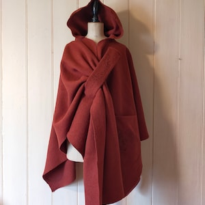May include: A rust-colored hooded cloak with a draped design and a front pocket. The cloak is displayed on a mannequin against a white-painted wood-paneled wall. The cloak has an embroidered design on the front.
