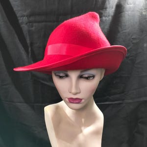 May include: A bright red hat with a wide brim and a ribbon detail. The hat features a distinctive, slightly pointed crown. The hat is displayed on a mannequin head with painted features.