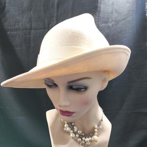 May include: A cream-coloured fedora hat with a wide brim and a ribbon band. The hat is displayed on a mannequin head, paired with a pearl necklace. The hat's design is classic and elegant.
