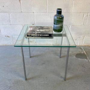 May include: A glass top coffee table with a chrome frame. A book titled "A History of Men's Fashion" by Michael  Kenna is on the table, along with a green and white swirl glass vase.