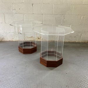 May include: Two clear acrylic side tables with a geometric design. Each table has a clear, octagonal top and a dark brown, octagonal base. The clear acrylic legs connect the top and base, creating a modern, minimalist aesthetic.
