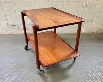 May include: A two-tiered wooden serving cart with a natural wood finish. The cart has four wheels and is made of solid wood.