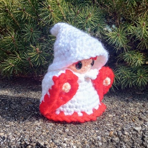 May include: A white crocheted gnome with red accents and large black eyes. The gnome is wearing a white hooded robe with red trim and red sleeves.