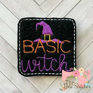 May include: A black felt patch with white stitching around the edges. The patch features a purple witch hat with orange and yellow accents and the text "Basic Witch" in orange, yellow, and purple thread.
