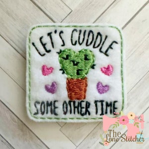 May include: White felt square with green embroidered cactus and the text "Let's cuddle some other time". There are pink and purple embroidered hearts around the cactus.