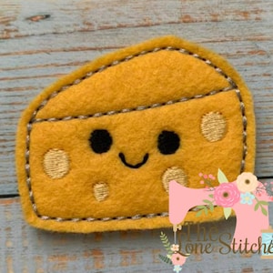May include: A yellow felt cheese with a smiling face and brown eyes. The cheese has a stitched border and small white dots.
