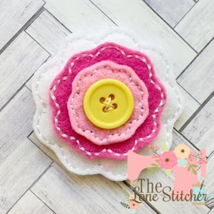 Puede incluir: Una flor de fieltro hecha a mano con capas de pétalos blancos, rosas y fucsias, cosida con hilo blanco. Un botón amarillo en el centro. Se ve el logotipo de The Lone Stitcher.