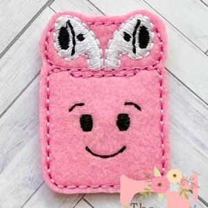 May include: Pink felt earphone holder with a smiling face and embroidered eyes. The holder has a pocket for earbuds and a stitched border.