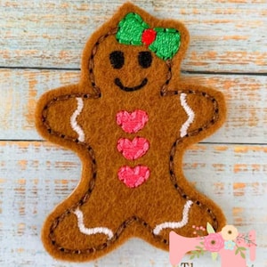 May include: A felt gingerbread person applique with a green bow and pink hearts. The gingerbread person is brown with white icing details.