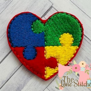 May include: A heart-shaped applique featuring a puzzle design. The puzzle pieces are in blue, green, and yellow, set against a red heart outline. The design is embroidered with a stitched texture.