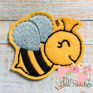 May include: A yellow and black embroidered bee with a light blue and white striped body. The bee is smiling and has a small, yellow heart on its head.