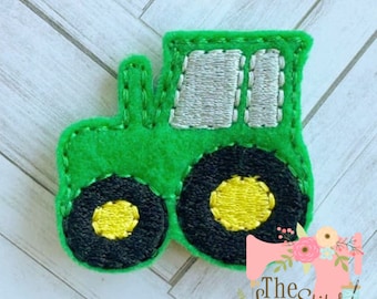 Può includere: Applique in feltro verde di un trattore con ruote nere e centri gialli. Il trattore ha una cabina grigia con dettagli di cucitura.