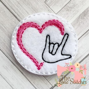 Könnte beinhalten: Weißer Filz-Kreis mit einem rosa Herzen und einem schwarzen Handzeichen für "Ich liebe dich" in Deutscher Gebärdensprache.