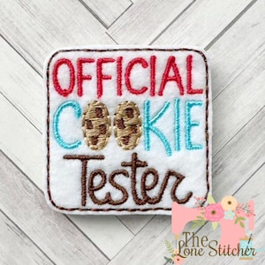 May include: White felt patch with brown stitching. The patch says "OFFICIAL COOKIE TESTER" in red, blue, and brown embroidery. Two brown chocolate chip cookies are embroidered in the center.