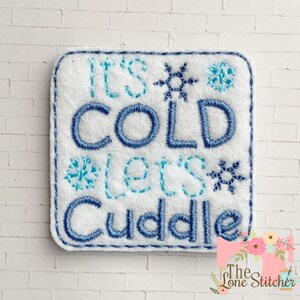 Puede incluir: Cuadrado de fieltro blanco con bordado azul que dice "Hace frío, abracemos" con diseños de copos de nieve.