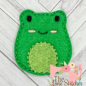May include: A green felt frog applique with a lighter green belly. The frog has black eyes and pink cheeks. The applique is stitched with green thread. The Lone Stitcher logo is in the bottom right corner.