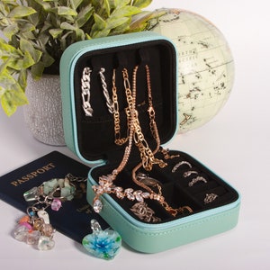 Puede incluir: Una caja de viaje para joyas de color azul claro con un interior negro. La caja está abierta y llena de varios collares, pulseras y pendientes. La caja está sobre una superficie blanca junto a un pasaporte y un globo terráqueo.