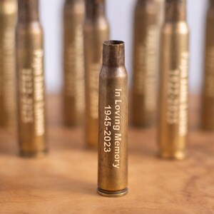 May include: A brass bullet casing engraved with "In Loving Memory 1945-2023" stands in front of several blurred casings. The casings are arranged on a wooden surface, creating a memorial or commemorative theme.