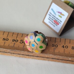 May include: A unique ring made from recycled colored pencils, featuring a cluster of colorful pencil tips. The ring includes a silver band and is accompanied by a small, triangular box with text that reads "This unique piece has been handmade from recycled colouring pencils."