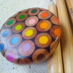Puede incluir: Un objeto redondo, colorido y con forma de mosaico, hecho con los extremos de lápices de colores afilados. El objeto está hecho de muchos colores diferentes, incluyendo rojo, naranja, amarillo, verde, azul y morado. El objeto está sobre una superficie marrón claro.