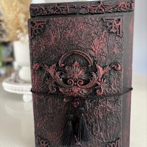 May include: A decorative journal in dark red and black. It features ornate, raised embellishments and a black tassel. The cover has a textured, crinkled appearance, and is bound with a black cord. The journal is suitable for writing.