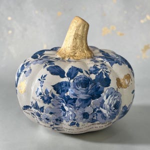 May include: A decorative pumpkin featuring a white base with a blue floral pattern and text snippets. The pumpkin has a textured gold-painted stem and gold leaf accents on the surface. The design includes various shades of blue roses and leaves.