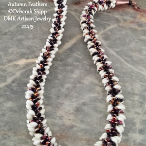Op de afbeelding: Een witte en zwarte kralenketting met koperen eindkappen. De kralen zijn in een chevron-patroon gerangschikt. De ketting heet "Autumn Feathers" en is van DMK Artisan Jewelry. Het artikelnummer is 22415.