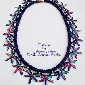 May include: A handcrafted necklace featuring a dark blue braided cord with iridescent flower-shaped beads. The necklace has a gold-colored clasp and is adorned with small gold beads. The text "Estrella by Deborah Shipp DMK Artisan Jewelry" is visible.