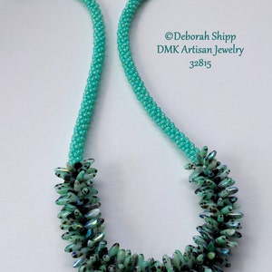 Puede incluir: Un collar de cuentas turquesa con un racimo de cuentas verdes y negras iridiscentes en el centro. El collar está hecho por DMK Artisan Jewelry y está etiquetado con el número 32815.