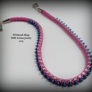 Pode incluir: Um colar de contas rosa e azul com fechos prateados. O colar é feito com contas pequenas e facetadas e tem um design geométrico. ©Deborah Shipp DMK Artisan Jewelry 21115