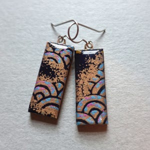 Könnte beinhalten: Ein Paar schwarze und goldene Ohrringe mit einem blauen und rosa Muster. Die Ohrringe sind rechteckig und haben einen silbernen Haken.