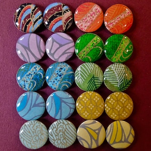 Könnte beinhalten: Ein Set mit 18 runden Knöpfen mit verschiedenen bunten Mustern und Designs. Die Knöpfe sind in einem Raster aus sechs Reihen und drei Spalten angeordnet. Die Muster umfassen geometrische Formen, florale Motive und abstrakte Designs.