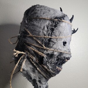 May include: A distressed, gray burlap mask with dark accents and twine detailing. The mask features a rough, torn texture and small, dark horns. The eyes are visible through small holes, and the mouth is slightly open, revealing teeth. A creepy Halloween decoration.