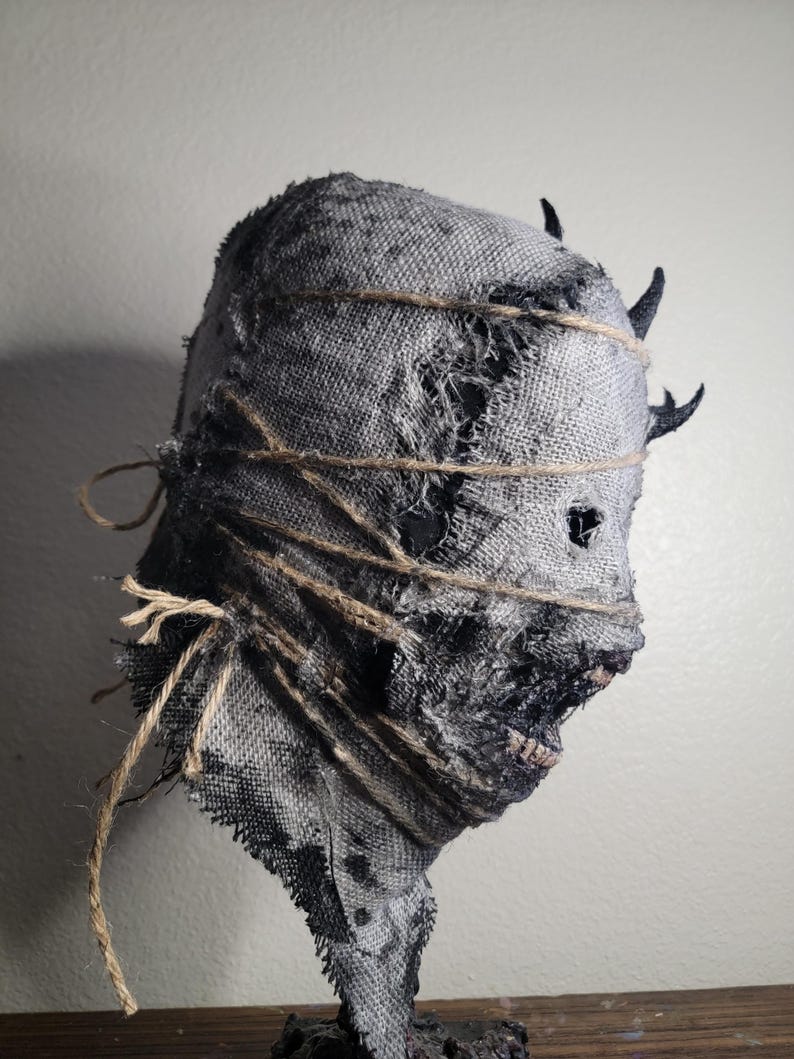 May include: A distressed, gray burlap mask with dark accents and twine detailing. The mask features a rough, torn texture and small, dark horns. The eyes are visible through small holes, and the mouth is slightly open, revealing teeth. A creepy Halloween decoration.