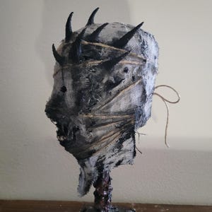 May include: A handmade, grotesque mask sculpture. The mask is primarily gray and black, with a textured, fabric-like appearance. It features several black, horn-like protrusions and is bound with twine. The base is dark and textured, suggesting a rough, organic material.