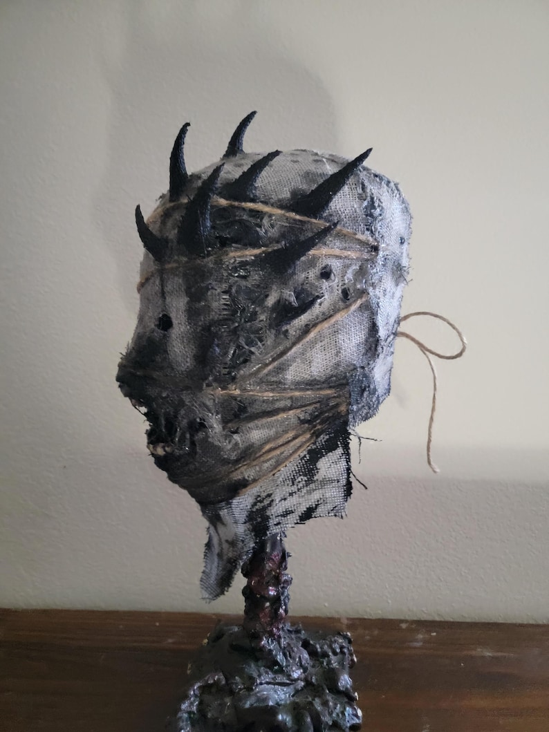May include: A handmade, grotesque mask sculpture. The mask is primarily gray and black, with a textured, fabric-like appearance. It features several black, horn-like protrusions and is bound with twine. The base is dark and textured, suggesting a rough, organic material.