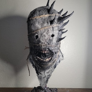 May include: A grotesque, handmade mask with a burlap texture, featuring jagged, dark spikes around the head. The mask has stitched details, eye holes, and a wide, menacing grin revealing teeth. It sits on a textured, dark base.