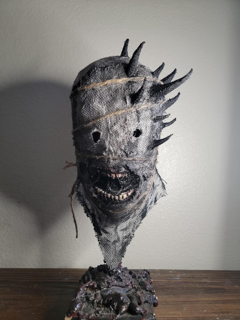 May include: A grotesque, handmade mask with a burlap texture, featuring jagged, dark spikes around the head. The mask has stitched details, eye holes, and a wide, menacing grin revealing teeth. It sits on a textured, dark base.