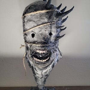 May include: A handmade, textured mask with a menacing appearance. The mask is primarily gray with black accents, featuring jagged horns and a stitched burlap design. The mouth is open, revealing teeth. The mask is mounted on a black base.