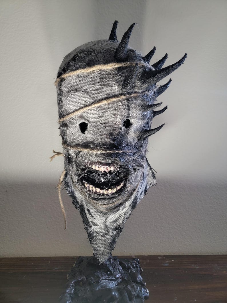 May include: A handmade, textured mask with a menacing appearance. The mask is primarily gray with black accents, featuring jagged horns and a stitched burlap design. The mouth is open, revealing teeth. The mask is mounted on a black base.