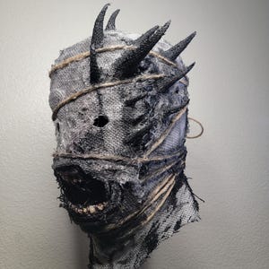 May include: A handmade, textured mask with a distressed appearance. The mask is primarily gray and black, with visible stitching and twine accents. It features multiple black horns and an open mouth revealing teeth, creating a menacing aesthetic. Ideal for Halloween or cosplay.