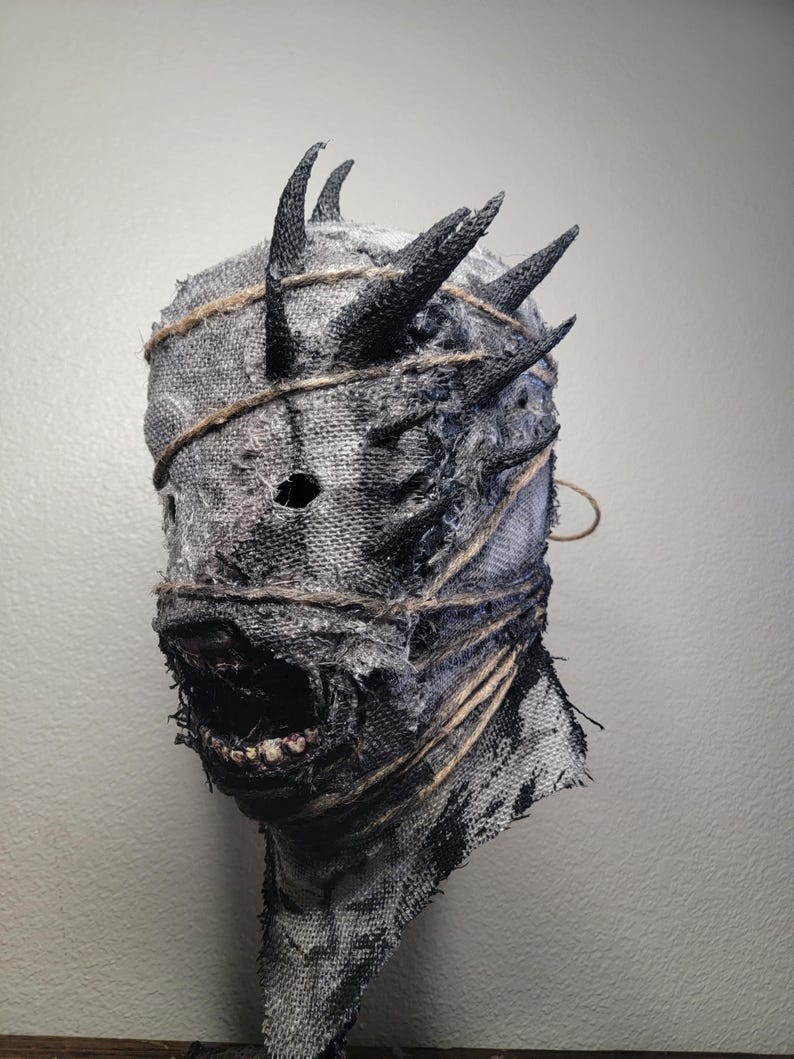 May include: A handmade, textured mask with a distressed appearance. The mask is primarily gray and black, with visible stitching and twine accents. It features multiple black horns and an open mouth revealing teeth, creating a menacing aesthetic. Ideal for Halloween or cosplay.